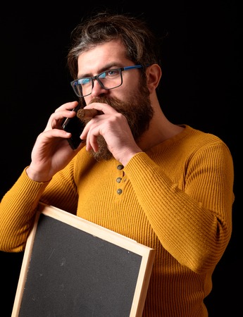 handsome bearded hipster man with long lush beard moustache and stylish hair on serious face in glasses yellow sweater holding board with cigar speaks by phone on black studio background, copy spaceの写真素材