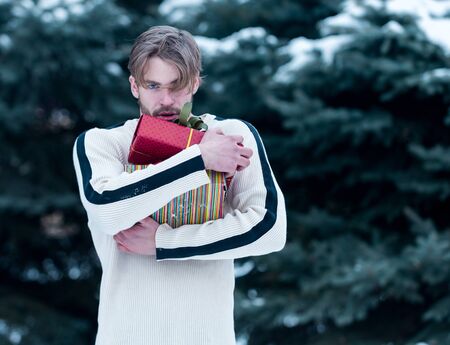 handsome bearded man or sexy guy in sweater in winter outdoor at green fir tree with snow holds present valentines box on natural background, copy spaceの写真素材