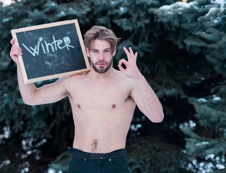 handsome bearded man or sexy guy with muscular body, chest and torso holds blackboard or chalkboard with winter text at green fir tree with snow, shows ok gesture on natural background, copy spaceの写真素材