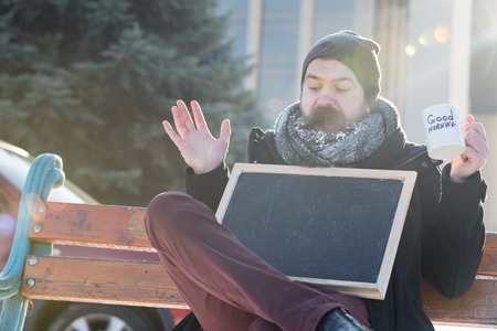 Excited man, bearded hipster with beard and moustache covered with white frost sits on wooden bench with blank black board or chalkboard and cup on snowy winter day on natural background, copy spaceの写真素材