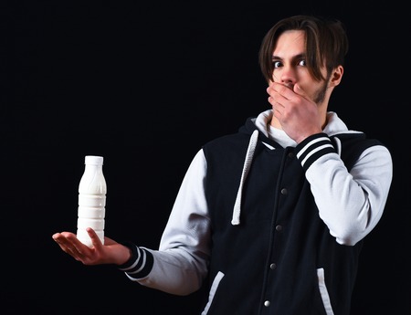 sportive handsome bearded guy in baseball jacket and surprised face holding kefir or yogurt in studio on black studio backgroundの写真素材