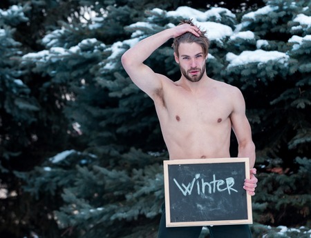 handsome bearded man or sexy guy with muscular body, chest and torso holds blackboard or chalkboard with winter text at green fir tree with snow on natural background, copy spaceの写真素材