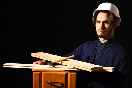 handsome bearded builder or foreman in blue sweater and helmet on serious face holding wooden bedside with boards or hummer on black studio backgroundの写真素材