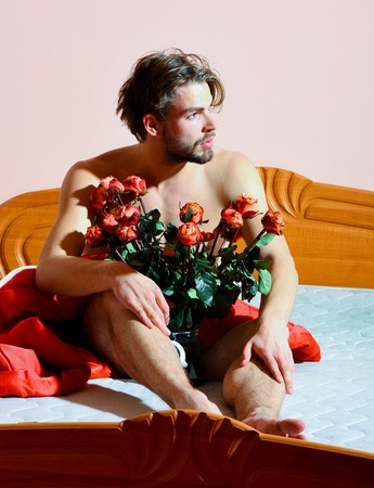 young handsome bearded sexy macho man with stylish beard sitting naked on bed under red blanket with bouquet of orange roses in transparent vaseの写真素材