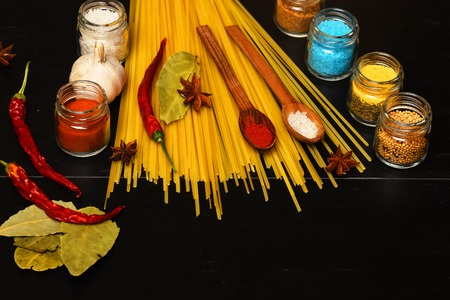 italian pasta with spice in jar, chili pepper, badian, bay leaf and garlic on black background as cooking products with wooden spoon, copy spaceの写真素材