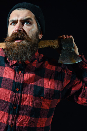 Handsome surprised man or lumberjack, bearded hipster, with beard and moustache in red checkered shirt keeps axe on shoulders isolated on black backgroundの写真素材