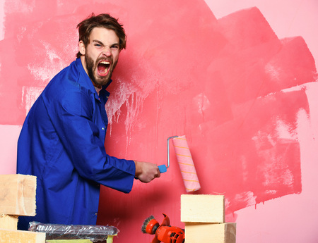 handsome bearded builder man with disheveled hair on shouting face in blue cloak and building tools on table painting wall with paint roller in studio on pink backgroundの写真素材