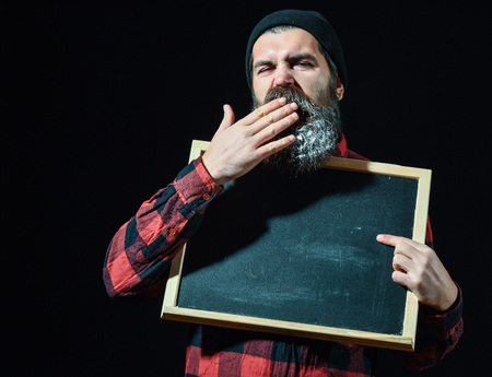 Bored man, bearded hipster with beard and moustache in red checkered shirt yawns with empty chalkboard or board isolated on black background, copy spaceの写真素材