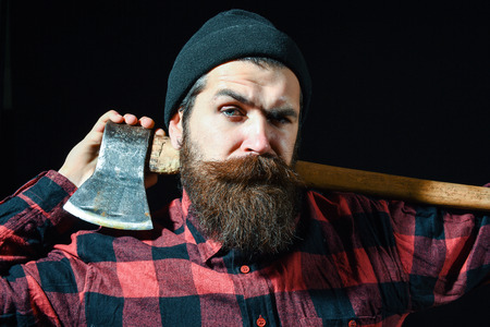 Handsome frown man or lumberjack, bearded hipster, with beard and moustache in red checkered shirt keeps axe on shoulders isolated on black backgroundの写真素材