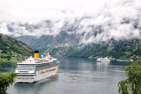 Norway - January 25, 2010: cruiser ship or liner in fjord, bay or harbor, calm water surrounded by mountains on rocky sea shore covered with green trees on summer day on cloudy white skyのeditorial素材