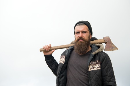 Handsome man hipster or guy with beard and moustache on serious face in hat and jacket holds rusty axe with wooden hilt outdoor against cloudy sky on natural background, copy spaceの写真素材