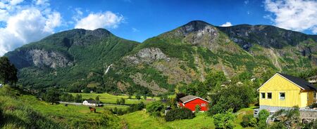 Picturesque village with small houses, cottages, on hilly rocky mountains covered with green trees and grass on sunny summer day on cloudy blue sky backgroundの写真素材