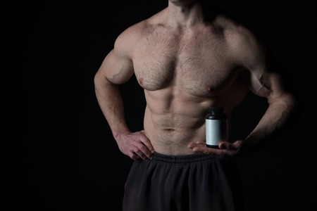 handsome macho man or sexy muscular guy with athlete body and torso holds white pill jar for drud, steroid, vitamin or doping on black backgroundの写真素材