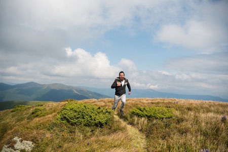 Handsome man hipster or guy runner with beard and moustache in hat run sunny outdoor on mountain top with green grass against cloudy sky on natural backgroundの写真素材