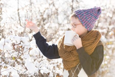 small boy or cute nerd kid in glasses, hat, sweater and fashionable knitted scarf holds cup and...