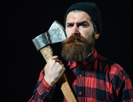 Handsome man or lumberjack, bearded hipster with beard and moustache in hat holds old rusty iron blade of axe isolated on black backgroundの写真素材