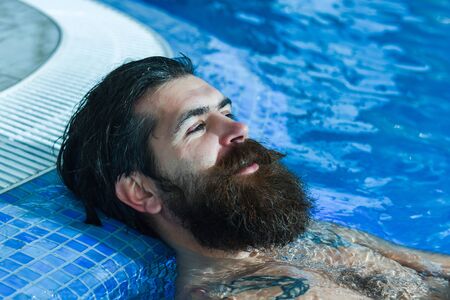 Handsome man or swimmer, bearded sexy hipster with wet beard, moustache and hair relaxes in pool indoors on blue water backgroundの写真素材