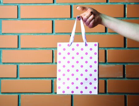 white color polka dot paper shopping bag or spotted package in female hand as holiday present or gift on brick wall background, copy spaceの写真素材