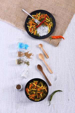 set of plates with pasta, chili pepper, spoon, fork, various spices in jars: salt, mustard, coriander, sackcloth napkin on white grey wooden background, top view, copy spaceの写真素材