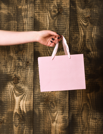 pink color paper shopping bag or textured package in female hand as holiday present or gift on wooden background, copy spaceの写真素材