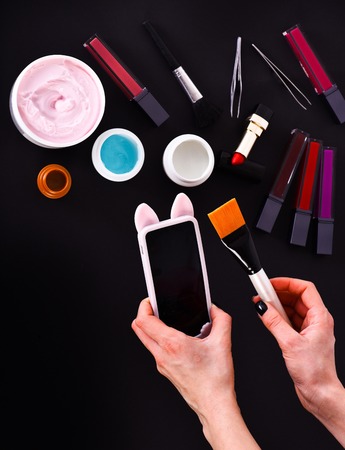 set of various cosmetics: cream, gel, lipsticks, lip gloss, brush, powder puff, tweezers and cell phone in hands isolated on black background, copy space, top viewの写真素材
