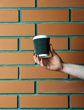 brown color paper or plastic cup for coffee drink as morning refreshment in female hand on brick wall background, copy spaceの写真素材