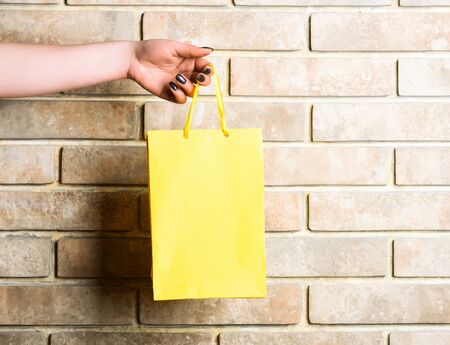 yellow color paper shopping bag or package in female hand as holiday present or gift on brick wall background, copy spaceの写真素材