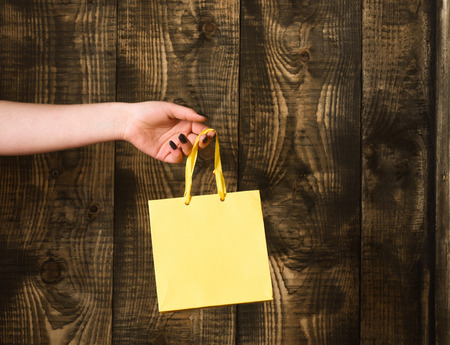yellow color paper shopping bag or package in female hand as holiday present or gift on wooden background, copy spaceの写真素材