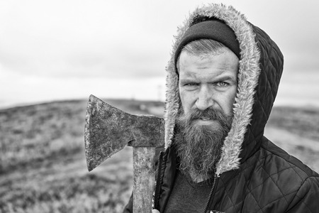 Handsome man hipster or guy with beard and moustache on serious face in hat and jacket holds rusty axe with wooden hilt outdoor on mountain top against cloudy sky on natural background, copy space, black and whiteの写真素材