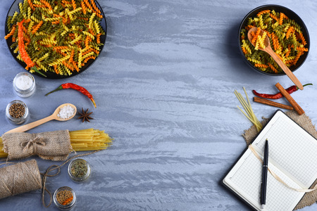 Colorful fusilli in two bowls, bunch of spaghetti in sackcloth, open notebook with pen, string bobbin, wooden spoons, red peppers, cinnamon sticks, anise, spices on grey background, copy spaceの写真素材