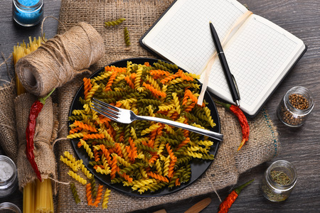 Fusilli pasta in bowl with fork and open notebook with pen, spaghetti bunch, string bobbin, red peppers, anise, spices, salt in jars and spoons on sackcloth on brown wooden background, copy spaceの写真素材
