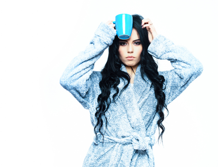 pretty serious cute sexy girl or beautiful woman with fashion makeup and curly long hair posing in blue velour bathrobe with cup isolated on white backgroundの写真素材