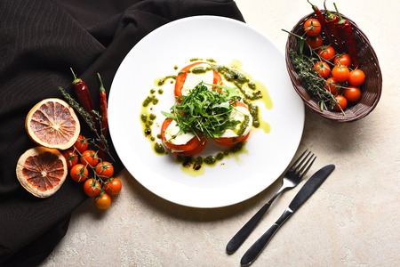 Tasty tomatoes sliced with mozzarella cheese and fresh green arugula salad on plate on black fabric and cherry, chilly peppers, spices, herbs in wicker basket, fork and knife on white backgroundの写真素材