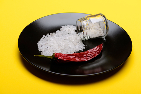 Glass jar with white salt crystals spilled and red chilly pepper on black plate for salting and seasoning food on yellow backgroundの写真素材
