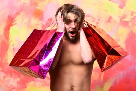 Caucasian surprised sexy macho man with moustache shows six packs and abs on muscle torso holding red packages or bags on colorful studio backgroundの写真素材