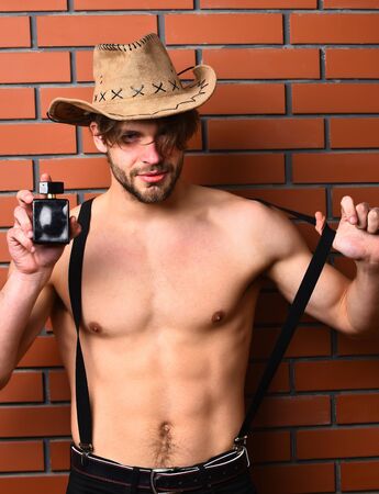 Caucasian macho man with moustache shows six packs on muscle torso in pants with suspenders and cowboy hat holding perfume on brown brick wall studio backgroundの写真素材