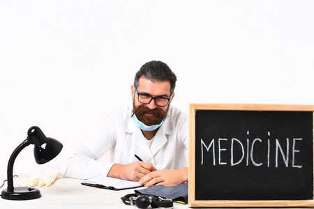 caucasian smiling happy doctor or unshaven hipster in mask, medical gown sitting at table with board "Medicine" inscription isolated on white studio backgroundの写真素材