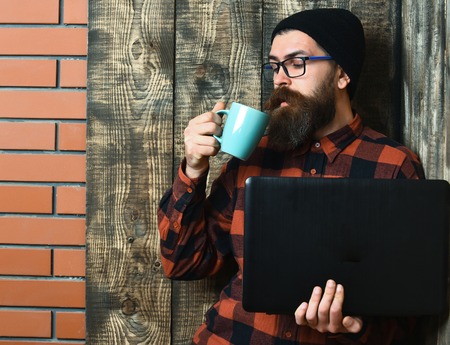 caucasian serious unshaven hipster holding laptop drink from mag or cup in red black checkered shirt with hat and glasses on brown vintage wooden studio backgroundの写真素材