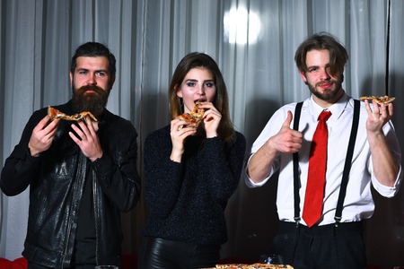Happy friends, pretty girl and two bearded men, hipsters with beards, smiling and eating tasty pizza slices at party in pizzeria, cafe or restaurant on white curtainの写真素材