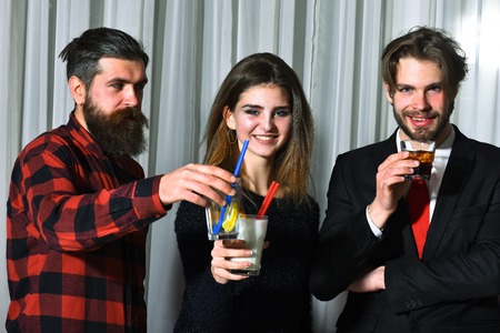 Happy friends, pretty girl and two bearded men, hipsters with beard, in red plaid shirt and business suit smiling clinking glasses with cocktails and alcohol drinks at party in cafe on white curtainの写真素材