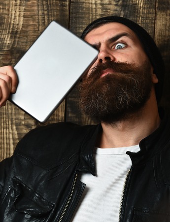 caucasian surprised unshaven hipster holding tablet in black leather jacket on brown vintage wooden studio backgroundの写真素材