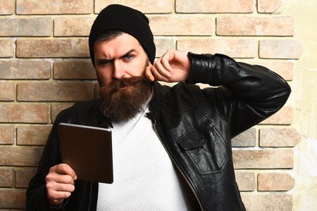 caucasian serious unshaven hipster holding tablet in black leather jacket on beige brick wall studio backgroundの写真素材