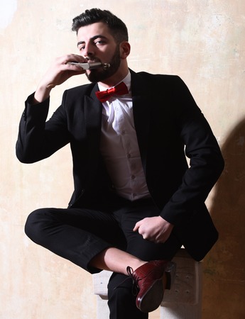 caucasian hipster with moustache in black elegant suit with bow-tie is sitting on chair, holding and drinking from flask with serious face on studio wall backgroundの写真素材