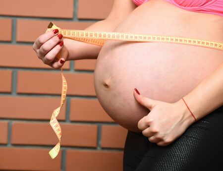female belly of pregnant woman with measuring tape on brown brick wall studio backgroundの写真素材