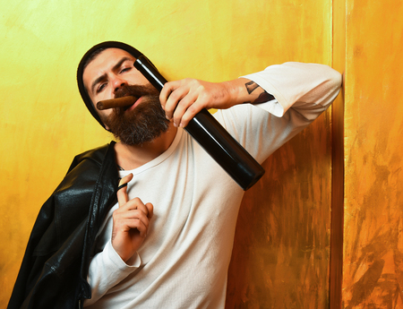 caucasian serious unshaven hipster holding bottle and smoking cigar with black leather jacket on shoulder, hat and white shirt on golden studio backgroundの写真素材