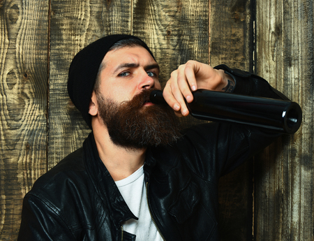 caucasian serious unshaven hipster holding drink from bottle with black leather jacket, hat and white shirt on brown vintage wooden studio backgroundの写真素材