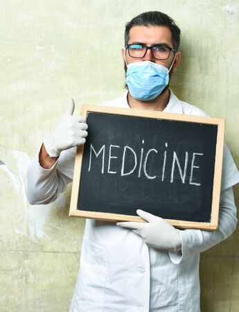 Bearded man, long beard. Brutal caucasian doctor or unshaven hipster, postgraduate student in medical gown, mask holding board with Medicine inscription on texture studio background. Medicine conceptの写真素材