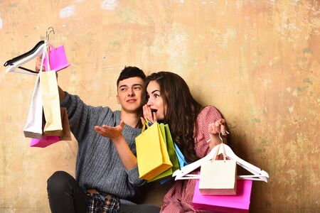 surprised happy couple of pretty sexy girl or cute fashionable woman with long brunette curly hair and young guy or handsome man holds hangers, colorful shopping bags or packages on beige backgroundの写真素材