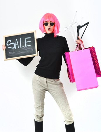cute fashionable sexy woman or pretty surprised girl in pink wig and fashion glasses holding colorful package or bags, hangers and board with sale inscription isolated on white studio backgroundの写真素材