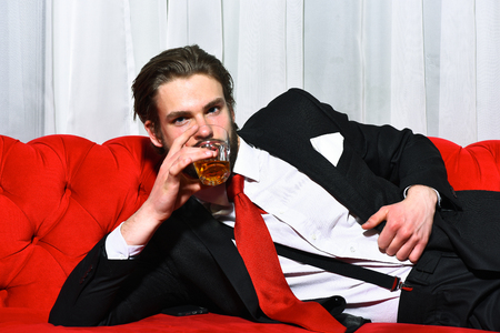 bearded man, businessman, long beard, brutal caucasian hipster with moustache holding glass with whiskey has serious face, unshaven guy with stylish hair in suit and red tie on sofaの写真素材